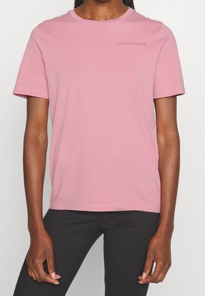 Person trägt ein schlichtes rosa kurzärmeliges T-Shirt mit dem "Peak Performance"-Logo auf der Brust und eine schwarze Hose und steht vor einem einfarbigen Hintergrund.