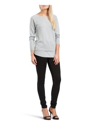 Pull en tricot gris clair avec un col rond, des manches longues, et un effet superposé sur un haut blanc, associé à un jean skinny noir et des talons noirs.