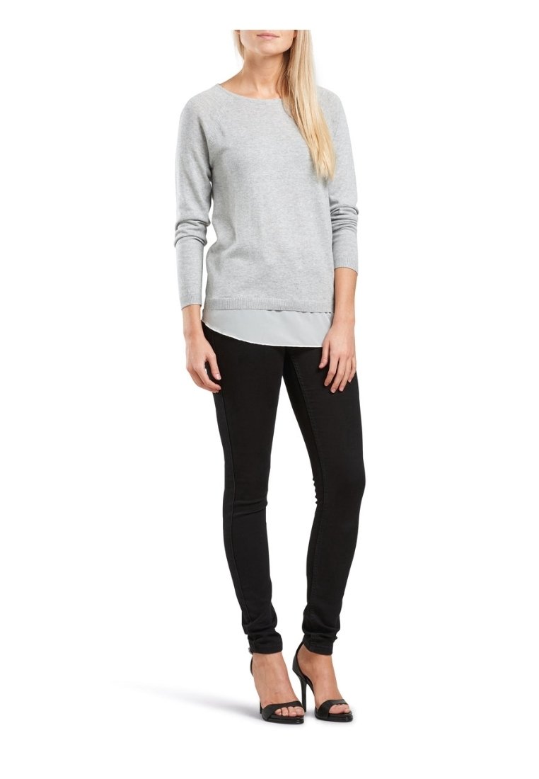 Pull en tricot gris clair avec un col rond, des manches longues, et un effet superposé sur un haut blanc, associé à un jean skinny noir et des talons noirs.