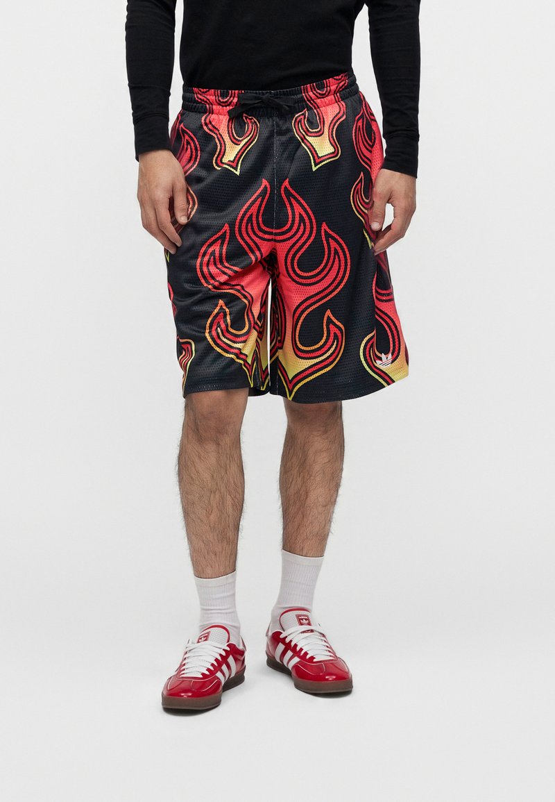 Uomo che indossa una maglietta nera a maniche lunghe e pantaloncini da basket neri con motivi a fiamma rossa e gialla, calzini bianchi e scarpe da ginnastica rosse.