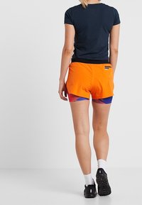 Kortärmad marinblå topp i kombination med livfulla orangea träningsshorts med ett blått och rött abstrakt mönster. Andningsbart material, avslappnad passform.