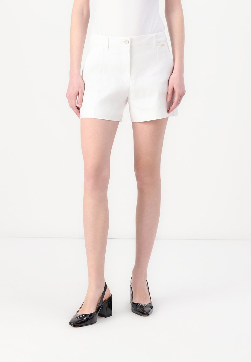 Hvite skreddersydde shorts har en subtil glans, sidelommer og frontknapp, og er kombinert med glinsende svarte hæler.