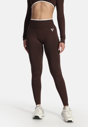 Donna che indossa leggings marroni a vita alta e crop top a maniche lunghe abbinato, con scarpe sportive bianche, in piedi su sfondo bianco.