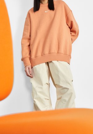 Personne portant un sweat-shirt Nike pêche oversize et un pantalon beige ample, debout entre deux chaises orange floues.