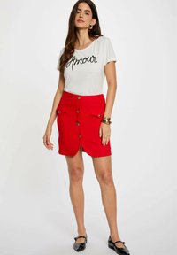 Rode rok met een sluiting aan de voorkant, twee zakken en een lichte split. Gecombineerd met een witte t-shirt met zwarte "Amour" tekst.