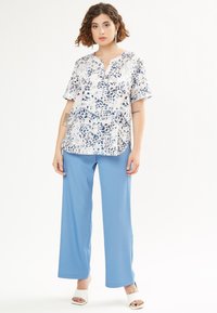 Blusa a fantasia floreale bianca e blu con maniche corte e scollatura a V, abbinata a pantaloni larghi azzurri e sandali bianchi con tacco blocco.