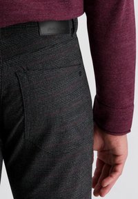 Grijze geblokte broek met subtiele rode lijnen, zwart leren label, gecombineerd met een bordeauxkleurige trui, waarbij de zak en de gestructureerde stof te zien zijn.