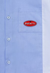 Camicia azzurra in cotone con bottoni. Presenta una tasca sul petto e un'etichetta ovale rossa con la scritta "BUGATTI" in bianco. Chiusura con due bottoni.