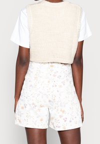 Persona vistiendo pantalones cortos de cintura alta con estampado floral blanco, una camiseta blanca y un chaleco sin mangas de punto beige, visto desde atrás.