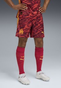 Czerwono-żółty strój piłkarski w wzory z pasującymi czerwonymi skarpetkami i białymi korkami. Zawiera logo Puma oraz logo Marsylii na szortach i skarpetkach.