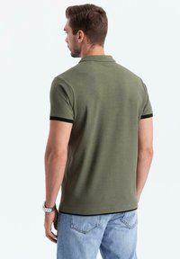 Camisa polo verde oliva con ribetes negros en las mangas, de manga corta y con un collar clásico. Combinada con unos vaqueros de mezclilla azul claro.
