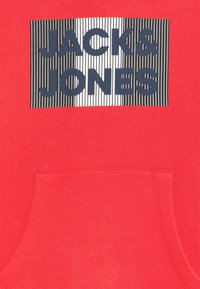 Sudadera roja con un gran gráfico rectangular que presenta el texto "JACK & JONES" en azul oscuro sobre un fondo de rayas en blanco y negro.