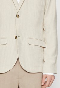 Beige linneblazer med två framfickor och en enkel knappstängning. Texturerat fiskbensmönster, slät tygfinish och rundade slag.