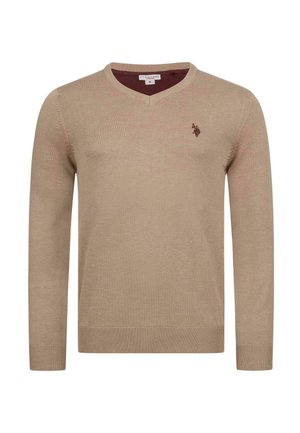 Mann mit dunklen Haaren, der einen beigen V-Ausschnittpullover mit kleinem gesticktem Logo auf der Brust und schwarze Jeans trägt, eine Hand in der Tasche.