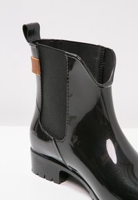 Botte de pluie noire brillante à la cheville avec panneaux latéraux élastiques, languette et semelle crantée épaisse sur fond blanc.