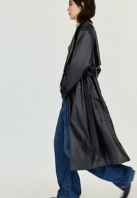 Femme marchant dans un long trench en cuir noir, un jean large bleu et des chaussures noires sur un fond clair neutre.