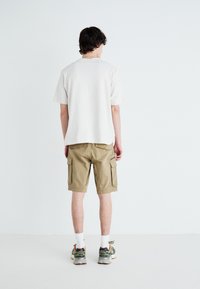 Khaki szorty cargo z bocznymi kieszeniami i jasnobeżowa bawełniana koszulka. Model ma na sobie sportowe sneakersy i białe skarpetki.