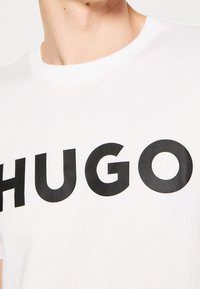 Zbliżenie na osobę noszącą białą koszulkę z dużymi czarnymi literami napisanymi "HUGO" na piersi.