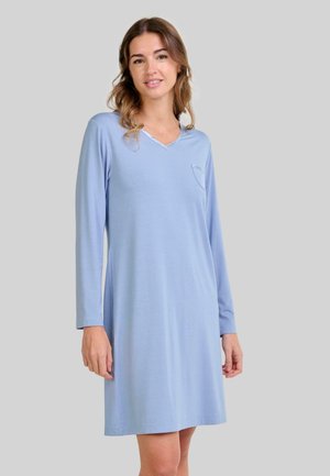 Mujer vestida con un vestido de manga larga azul claro, de largo hasta la rodilla, con un pequeño bolsillo en el pecho, de pie frente a un fondo gris liso.
