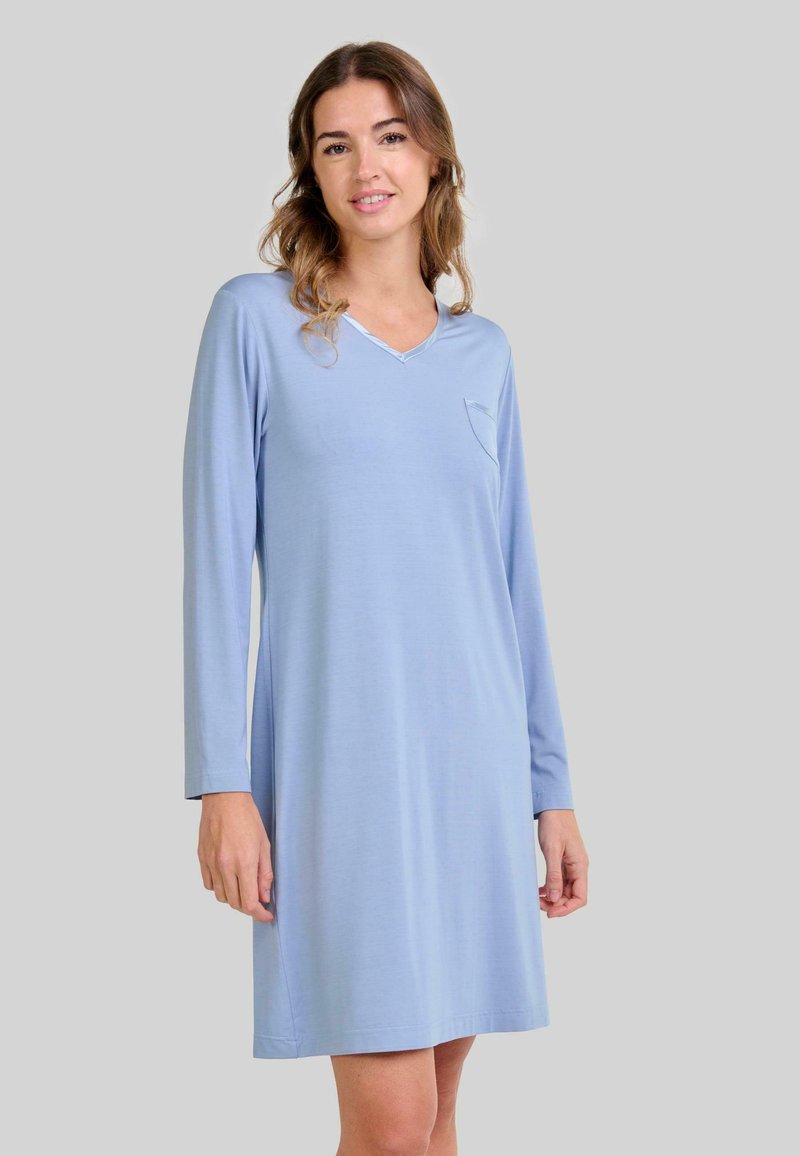 Mujer vestida con un vestido de manga larga azul claro, de largo hasta la rodilla, con un pequeño bolsillo en el pecho, de pie frente a un fondo gris liso.