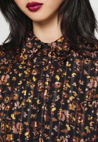 Femme portant un chemisier boutonné à motifs floraux sombres avec un col pointu et un rouge à lèvres rouge foncé, montrée du cou à la poitrine.