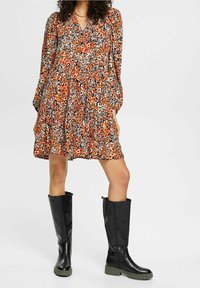 Robe fleurie à manches longues en orange, blanc et noir, cintrée à la taille. Associée à des bottes noires montantes avec des semelles vertes.