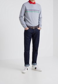 Maglione grigio con strisce orizzontali multicolori, camicia rossa con colletto sotto, jeans in denim scuro, cintura con motivo, scarpe bianche con dettagli colorati.