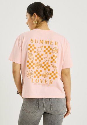 Mujer con cabello oscuro recogido en un moño lleva una camiseta rosa con gráficos naranjas de conchas marinas y estrellas de mar y texto «SUMMER LOVER SEA COLLECTION» en la espalda.