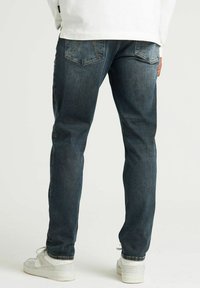 Jeans din denim albastru închis, cu croială ajustată, dotate cu buzunare decorative la spate și ușoare decolorări pe coapse. Purtate cu adidași albi.