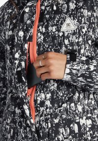 Black and white patterned jacket with coral zipper, reinforced seams, and triangular logo patch, featuring a chest pocket for storage.