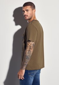 Olivově zelené bavlněné tričko s krátkým rukávem, na přední straně s grafickým designem. Nosené s modrými džíny. Tetování viditelné na paži.