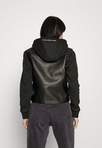 Černá a tmavě zelená bunda s kapucí, které mají kožený zadní díl a měkké látkové rukávy. Na kapuci je logo "Calvin Klein Jeans". Šedé kalhoty.