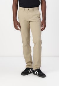 Pantalons en coton beige avec une coupe droite, dotés d'une fermeture à bouton à l'avant et de poches latérales, associés à des baskets noires.