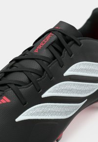 Μαύρο ποδοσφαιρικό παπούτσι με υφή, με κόκκινο λογότυπο «PREDATOR» και γκρι συνθετικές ρίγες. Διαθέτει μαύρα κορδόνια και λεπτομέρειες.