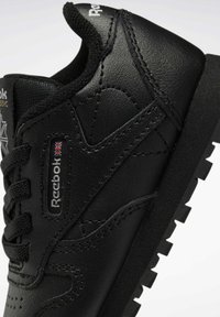 Svarta sneakers med en strukturerad läderöverdel, rund tå och en gummisula med ett robust mönster. Reebok-logotyp på sidan.