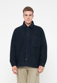 Νεαρός άνδρας ντύνεται με σκούρο ναυτικό κουμπωτό φλίς μπουφάν με τσέπη στήθους και μπεζ cargo παντελόνι, στέκεται μπροστά σε λευκό φόντο.