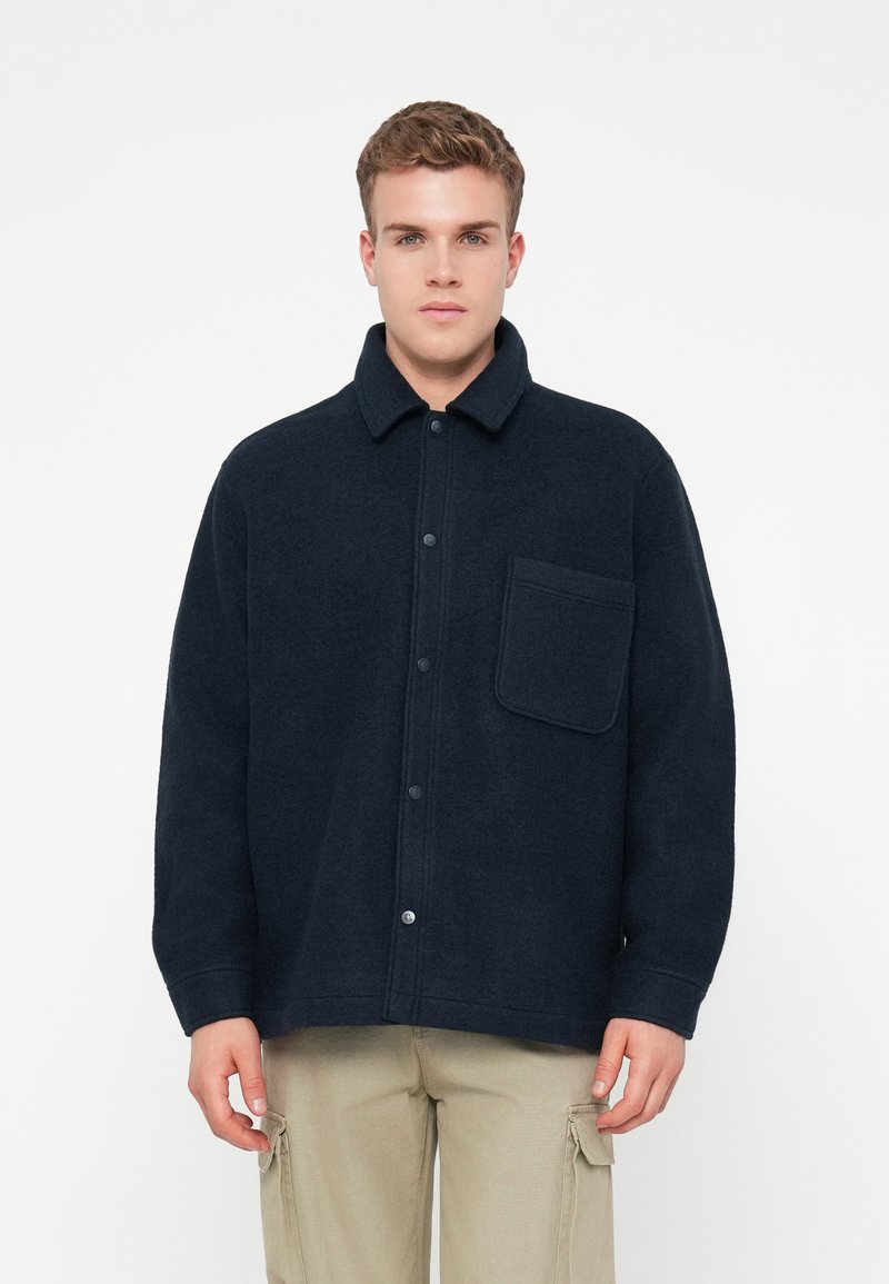 Νεαρός άνδρας ντύνεται με σκούρο ναυτικό κουμπωτό φλίς μπουφάν με τσέπη στήθους και μπεζ cargo παντελόνι, στέκεται μπροστά σε λευκό φόντο.