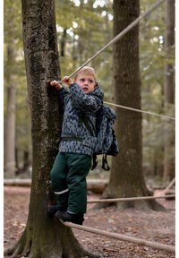 Barn i en mønstret grå og sort jakke og grønne bukser kravler op i et træ og holder fast i et reb. En rygsæk hviler på deres ryg.