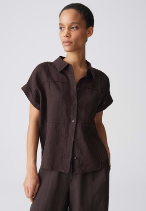 Vrouw draagt een losse donkerbruine korte mouwen blouse met knopen en bijpassende broek, kijkt opzij tegen een effen grijze achtergrond.