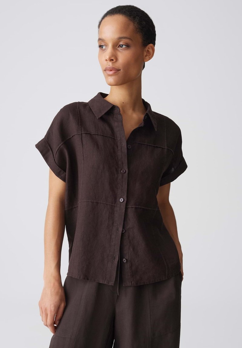 Femme portant une chemise ample marron foncé à manches courtes avec boutons et pantalon assorti, regardant sur le côté devant un fond gris uni.