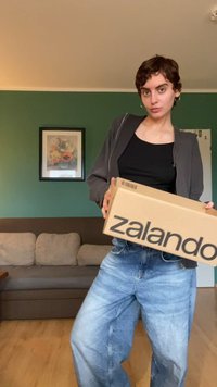 Hnědá krabice Zalando držená v rukou, s výrazným černým logem. Modelka má na sobě černý top bez rukávů, šedou bundu na zip a světlé modré široké džíny.