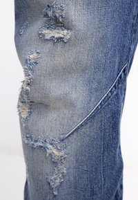 Jeans de denim de color azul claro con múltiples áreas deshilachadas y rasgadas a lo largo de la sección del muslo que muestran hilos blancos debajo.