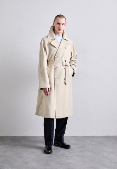 Trench coat beige doppiopetto con vita cintrata, ampi rever e texture morbida. Modello abbinato a pantaloni scuri e scarpe nere.