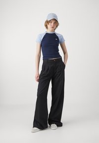 Haut à manches courtes bleu marine et clair associé à un pantalon large noir ; le mannequin porte une casquette bleu clair et des chaussures à enfiler beiges.