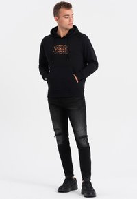 Felpa nera con cappuccio e tasca a marsupio, con una stampa grafica arancione sul petto. Abbinata a jeans scuri strappati e sneaker nere.
