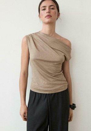 Femme portant un haut beige asymétrique sans manches, décolleté épaule dégagée, et un pantalon noir ample, se tenant devant un fond blanc uni.