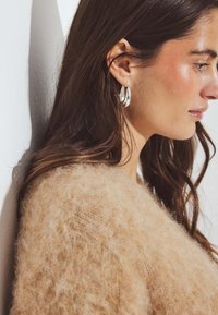 Sidabriniai žiediniai auskarai greta švelnaus smėlio spalvos pūkuoto megztinio, pabrėžiant megztinio tekstūrą ir auskarų blizgesį.