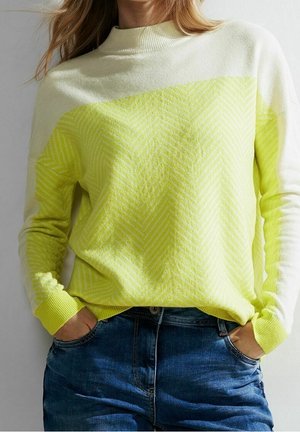 Person iført en gul og hvid gradient sweater med chevronmønster, stoppet ned i blå jeans med hænderne i lommerne.