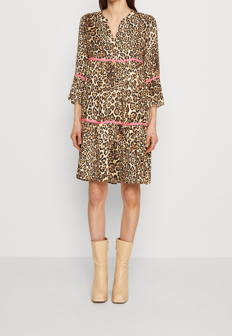 Femme portant une robe léopard longueur genou avec liseré rose et des bottines beiges, debout devant un fond clair et uni.