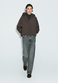 Donna che cammina indossando un giubbotto cappuccio oversize marrone scuro, dolcevita nera, jeans grigi larghi, cintura borchiata e scarpe marroni scure su sfondo neutro.